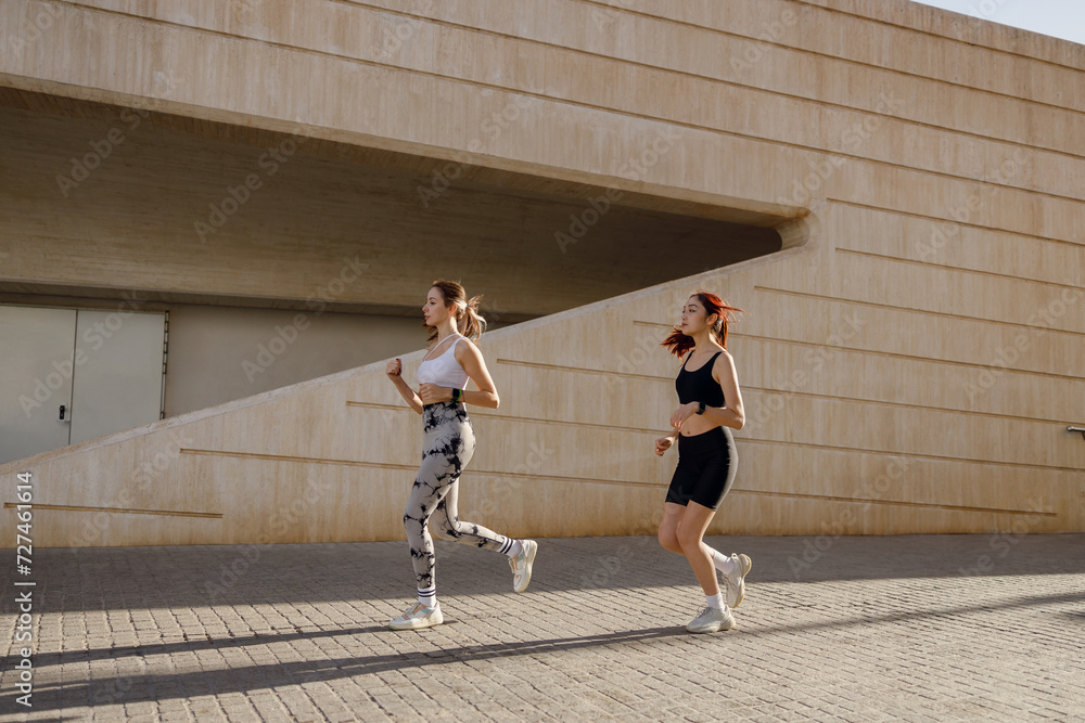 Obraz premium Two young women in sportswear are running on modern buildings background. Active lifestyle concept