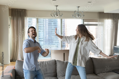 Millennial couple, excited man and woman having fun together, dancing in living room, sing song, move to favourite music celebrate relocation day, enjoy pastime active leisure in skyscraper apartment