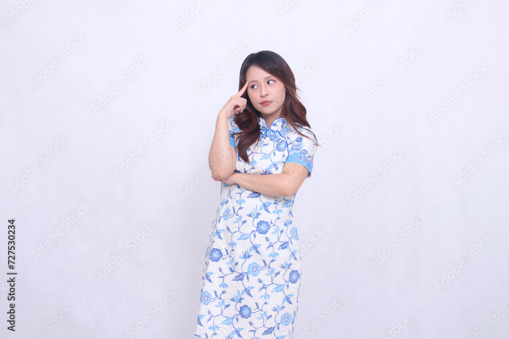 Beautiful Chinese Javanese woman frowning while thinking, wearing a modern Chinese dress, arms crossed, pointing to her forehead on a white background. happy Chinese New Year Beautiful Chinese Javanese woman frowning while thinking, wearing a modern Chinese dress, arms crossed, pointing to her forehead on a white background. happy Chinese New Year