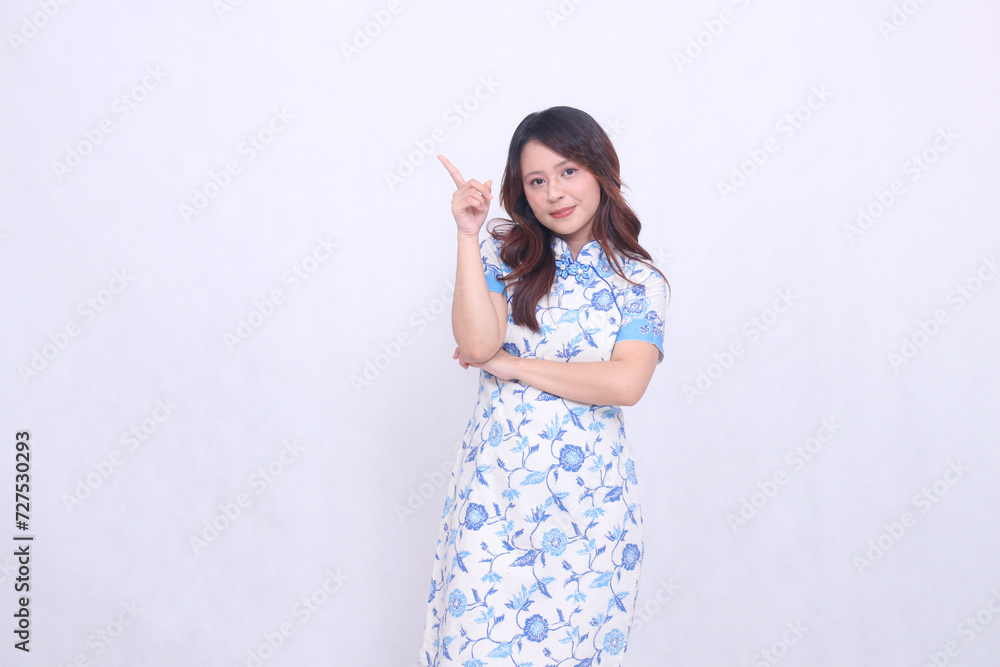 Beautiful Chinese Javanese woman smiling wearing a modern Chinese dress with her arms crossed pointing at the top left on a white background. happy Chinese New Year Beautiful Chinese Javanese woman smiling wearing a modern Chinese dress with her arms crossed pointing at the top left on a white background. happy Chinese New Year