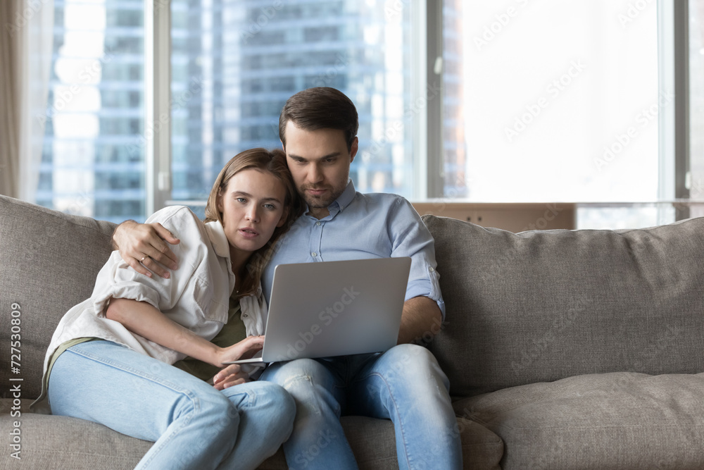 Millennial loving couple buying on internet, spend time using laptop resting on cozy sofa in modern living room, choose gifts, buy tickets, booking hotels, planning vacation. E-services clients, tech