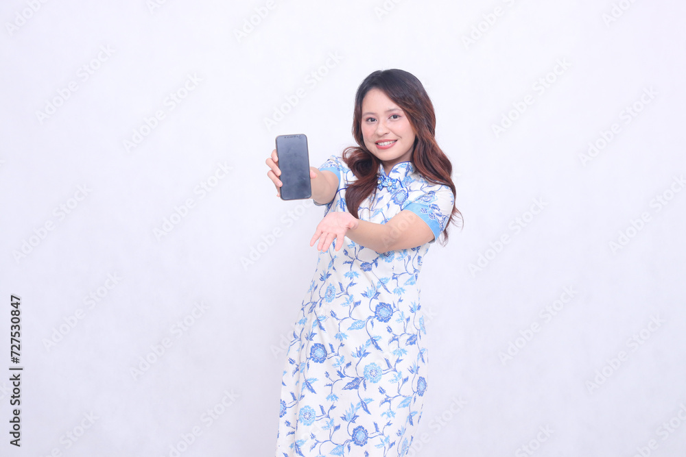 young Indonesian Javanese girl wearing a blue traditional Chinese dress cheerfully holding a gadget showing her cellphone screen raised to the front isolated on a white background young Indonesian Javanese girl wearing a blue traditional Chinese dress cheerfully holding a gadget showing her cellphone screen raised to the front isolated on a white background