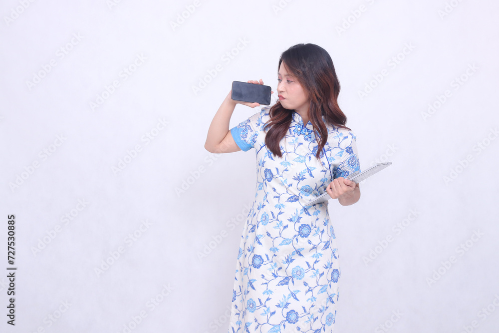 beautiful asian woman wearing modern chinese blue dress looking at cell phone frowning holding tablet and looking at cell phone screen isolated white background beautiful asian woman wearing modern chinese blue dress looking at cell phone frowning holding tablet and looking at cell phone screen isolated white background