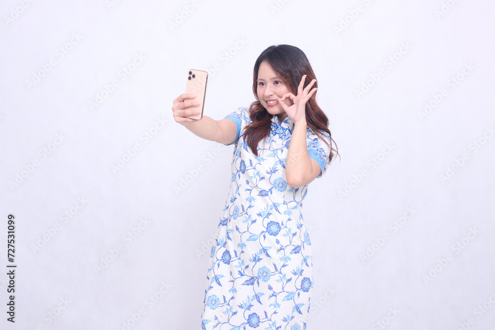 young asian javanese chinese woman smiling laughing agreeing wearing blue chinese dress looking at cellphone and operating it isolated white background young asian javanese chinese woman smiling laughing agreeing wearing blue chinese dress looking at cellphone and operating it isolated white background