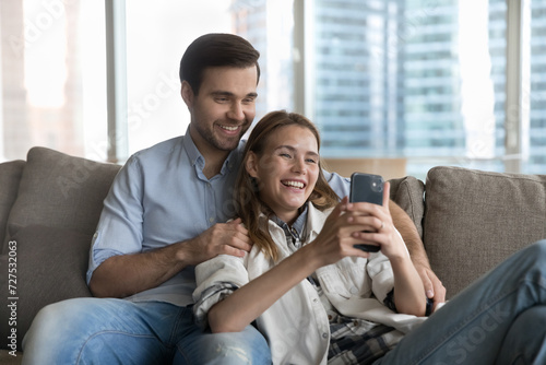 Beautiful happy millennial loving couple relaxing on sofa staring at cellphone screen watch funny videos, enjoy videoconference call to family via mobile videocall app, spend leisure using modern tech