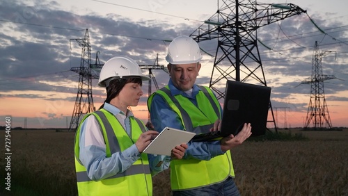 Engineers exchange digital data via computers near power transmission lines in sunset meadow. Man and woman electricians integrate software for power distribution substation improvement at twilight