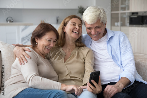 Excited happy older parents and adult daughter woman using media app on mobile phone together, watching funny content, taking group selfie, hugging, laughing, having fun