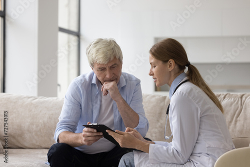 Serious doctor woman in white coat giving treatment recommendation to older man, explaining medical checkup results, diagnosis, therapy instructions, meeting with patient at home