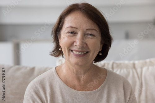 Positive mature older retired woman home head shot portrait. Female pensioner sitting on sofa, looking at camera with happy smile, enjoying retirement, talking on video conference call photo