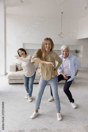 Happy excited adult daughter and joyful active senior parents dancing, looking at camera, smiling, laughing, having fun. Trainer training elder couple at home, enjoying activity to music in apartment