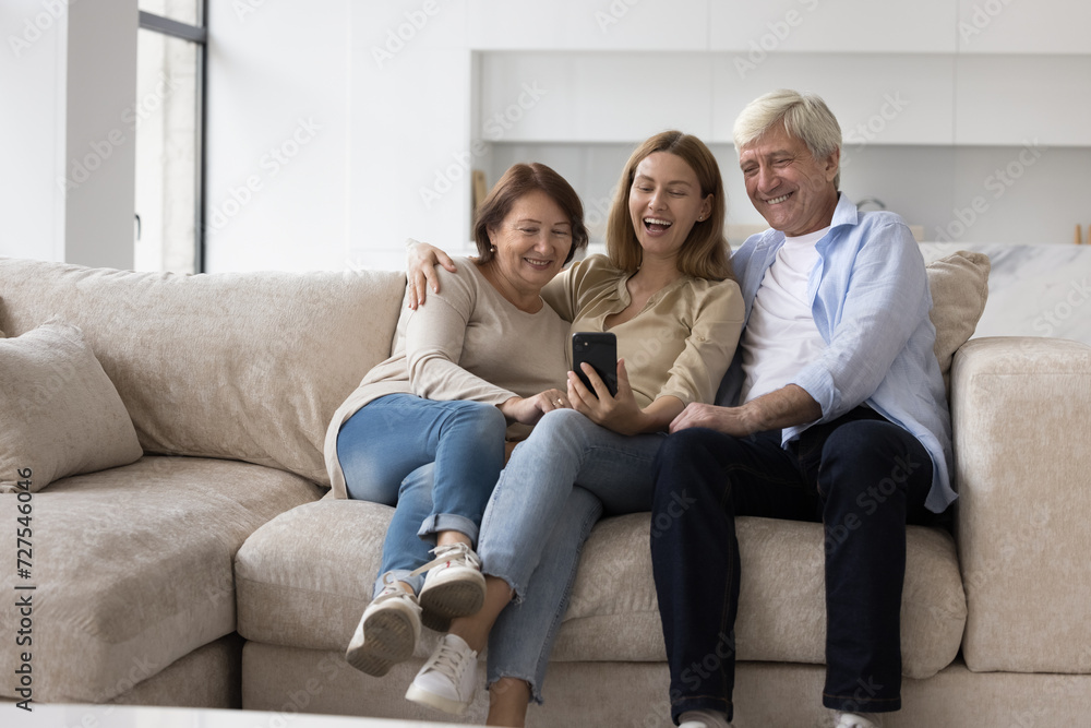 Cheerful elder couple of parents and adult child woman using smartphone for family video call, enjoying online conference conversation, talking selfie, laughing, posing, resting on home sofa