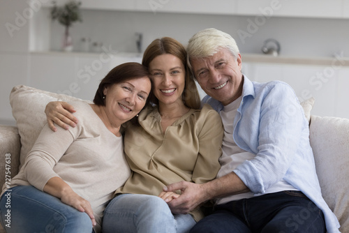 Happy adult daughter woman enjoying meeting with senior parents at home, hugging mature mom and dad with love, care, sitting close on couch, smiling, laughing, looking at camera. Family portrait