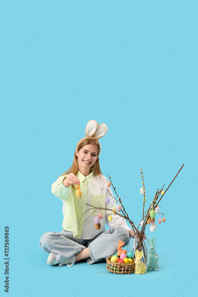 Young woman in bunny ears with Easter eggs and willow branches sitting on blue background