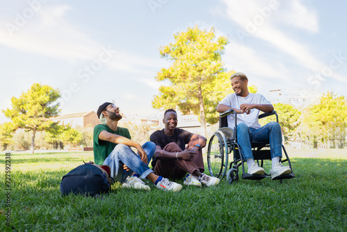 Young Black man in a wheelchair and friends laughing and having fun sitting on the grass in a park. Copy space.
