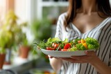 A woman is happily eating a healthy salad in a bright kitchen, surrounded by fresh vegetables, creating a nutritious and delicious meal