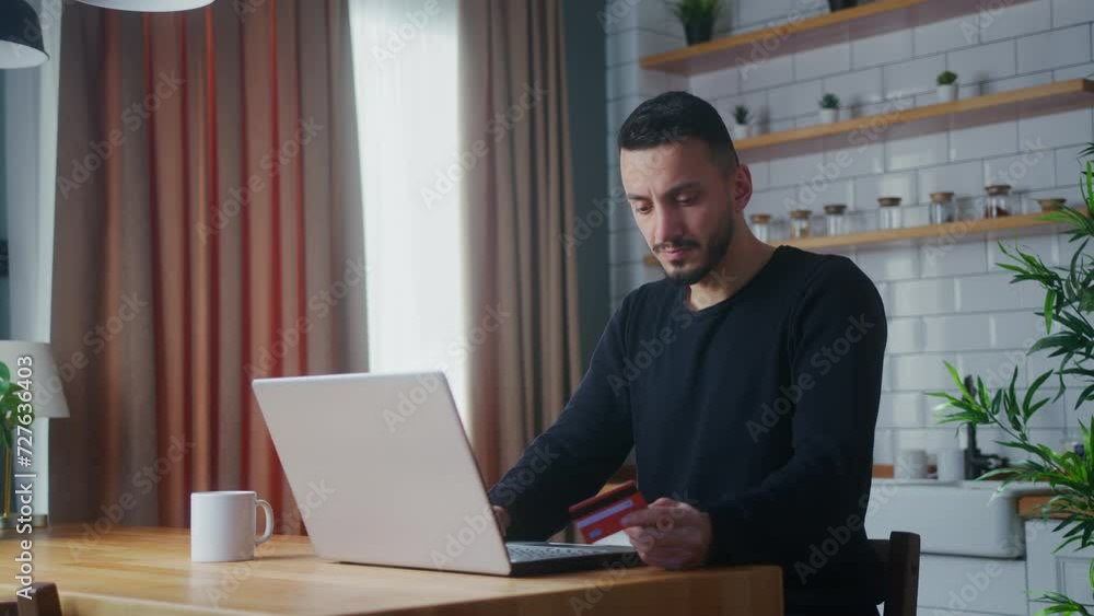 Adult man enter credit card number on laptop for shopping online, makes secure easy distant electronic payment while sitting in kitchen at home Adult man enter credit card number on laptop for shopping online, makes secure easy distant electronic payment while sitting in kitchen at home