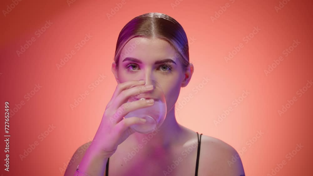 Young nordic woman drinks glass of cold water looking at camera, close up portrait shot in studio with orange background Young nordic woman drinks glass of cold water looking at camera, close up portrait shot in studio with orange background