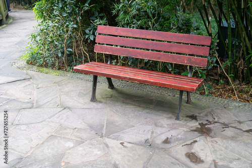 A maroon park bench made of wood and metal placed at the side of a concrete pathway inside a public conservation facility.