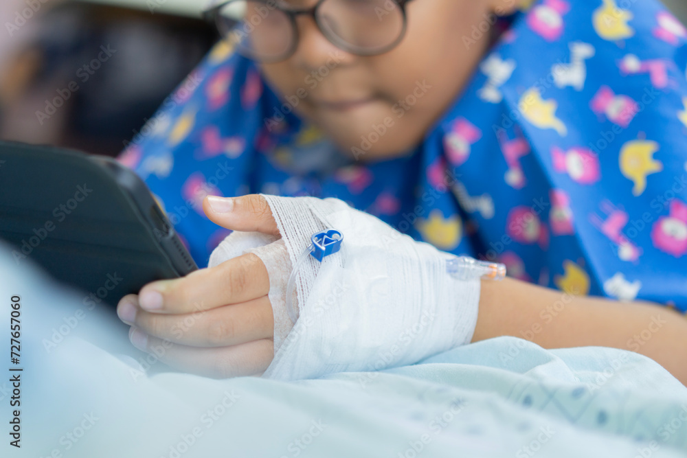Sick asian little child boy who have IV solution bandaged playing digital tablet to relax while staying in the hospital.