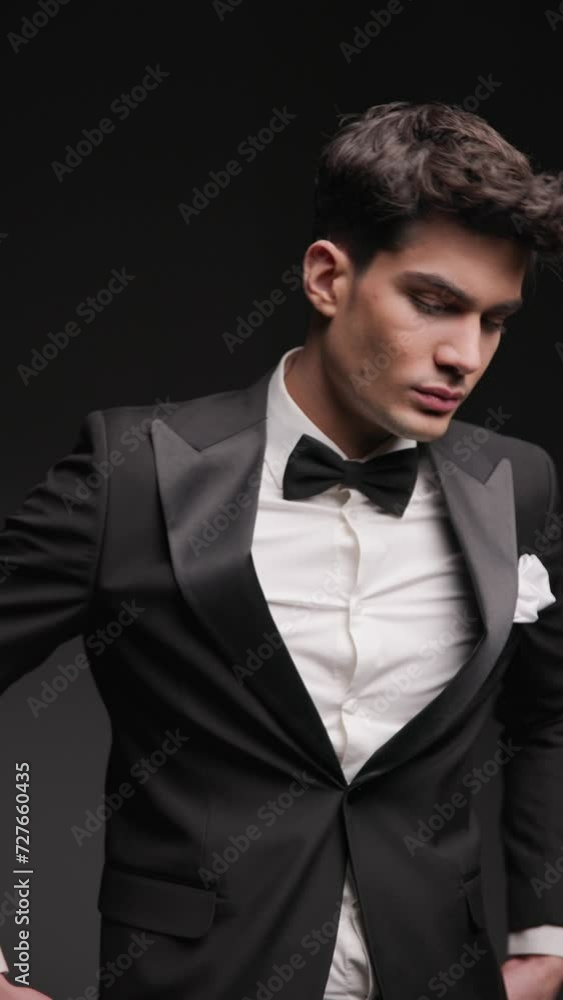 handsome elegant groom wearing black tuxedo, holding hands in pockets, looking down and fixing tux in front of grey background with flash lights on handsome elegant groom wearing black tuxedo, holding hands in pockets, looking down and fixing tux in front of grey background with flash lights on