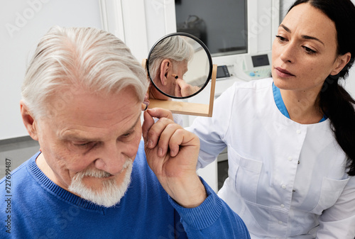 Senior man trying on hearing aid in front of mirror with female audiologist, fitting hearing device into his ear while audiology consultation photo