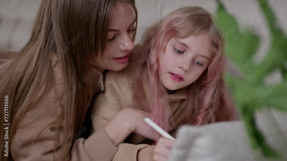 A young woman watching a rose-haired girl using a tablet with a stylus pen while lying on a couch in a room. A young woman watching a rose-haired girl using a tablet with a stylus pen while lying on a couch in a room.
