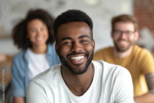A multiracial man smiling at the camera with his White and Hispanic coworkers are talking behind. 