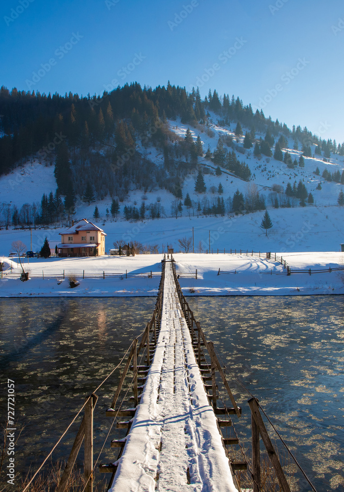 A narrow suspension bridge over a river in a mountain area in winter