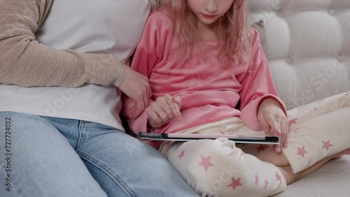 A cropped rose-haired girl using a tablet while sitting cross-legged on a light sofa by a young woman.