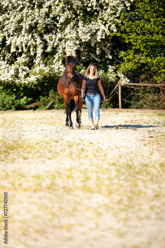 Young woman with a black shirt and short highlighted hair stands with her horse on a sunny riding arena.