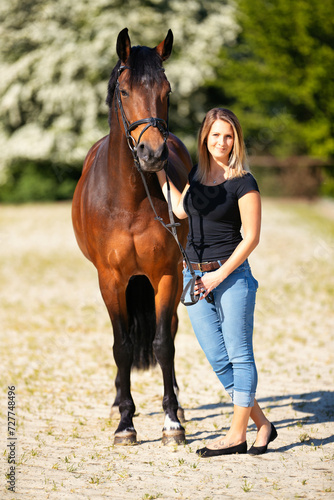 Young woman with a black shirt and short highlighted hair stands with her horse on a sunny riding arena.