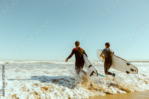 Photography Back view of group of friends with surfboards in wetsuit entering towards ocean