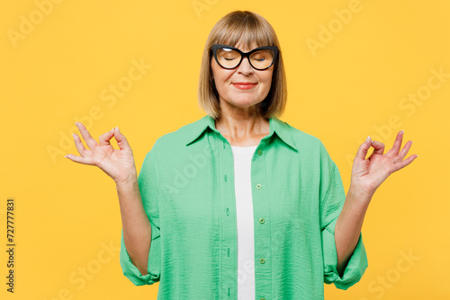 Elderly blonde woman 50s years old wear green shirt glasses casual clothes hold spread hands in yoga om aum gesture relax meditate try calm down isolated on plain yellow background. Lifestyle concept. photo