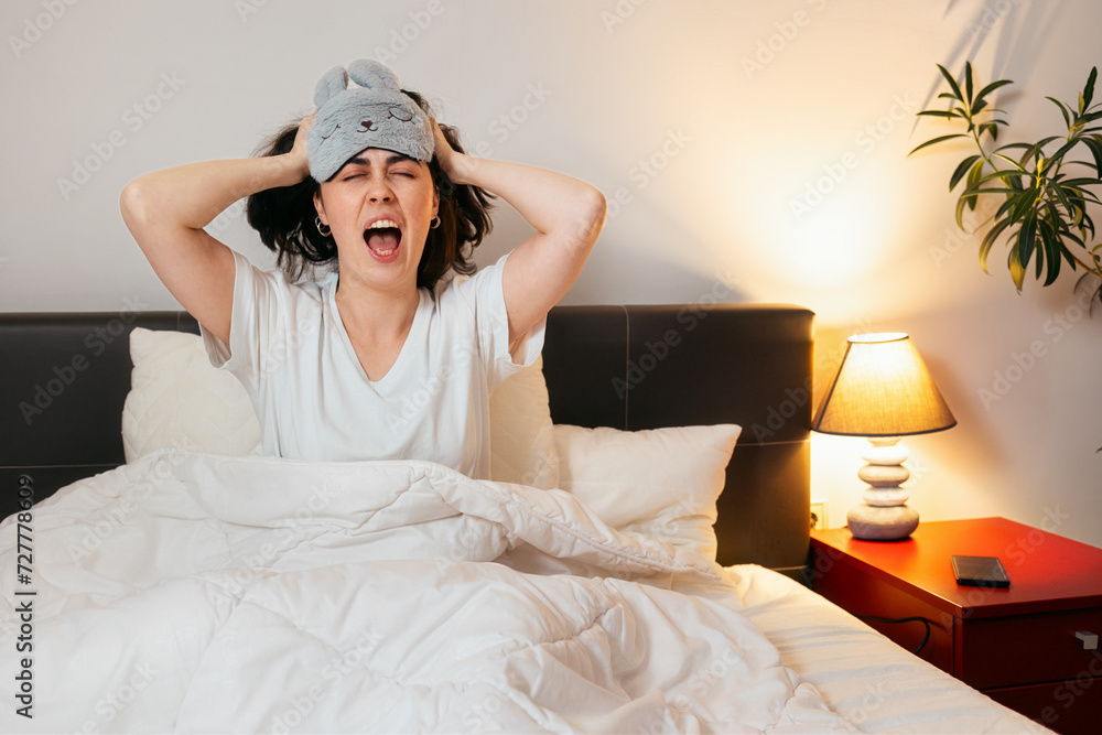 Irritated Caucasian woman with sleep mask shouts holds hands on head is sitting in bed. Insomnia due to noisy neighbors Irritated Caucasian woman with sleep mask shouts holds hands on head is sitting in bed. Insomnia due to noisy neighbors