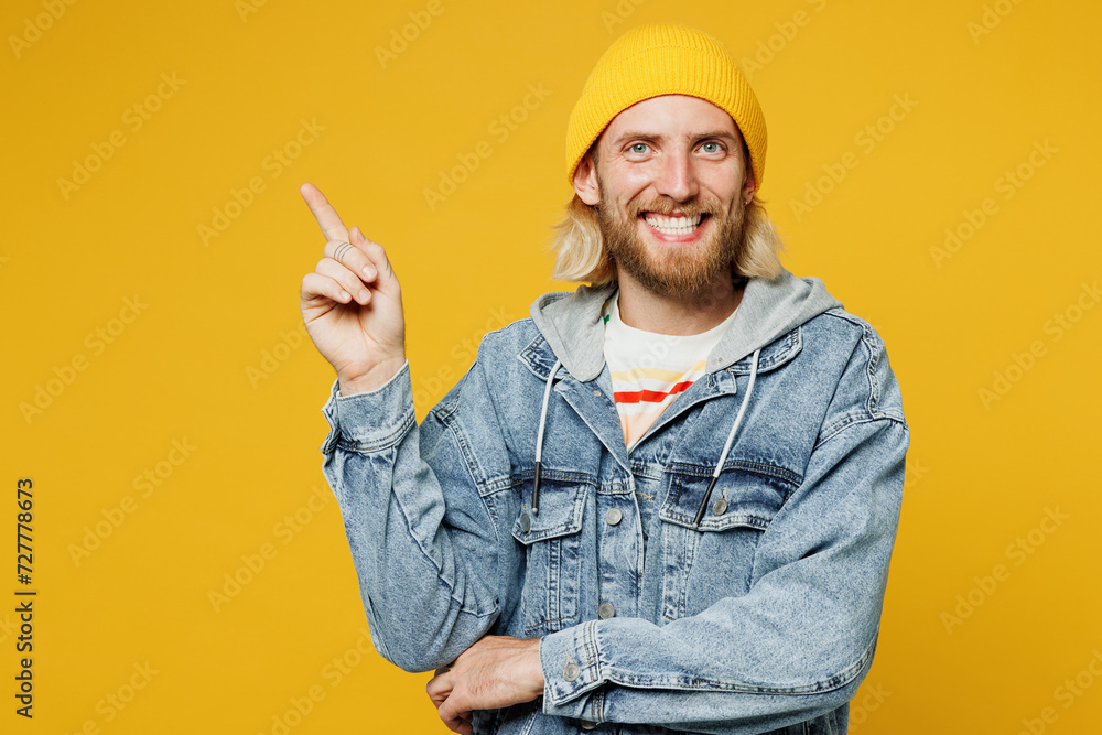 Young smiling happy fun blond man he wears denim shirt hoody beanie hat casual clothes point index finger aside on area mockup isolated on plain yellow background studio portrait. Lifestyle concept.