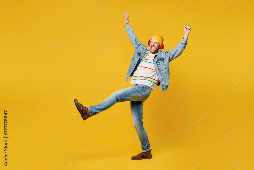 Full body young happy man he wear denim shirt hoody beanie hat casual clothes listen to music in headphones with raised up hands isolated on plain yellow background studio portrait. Lifestyle concept