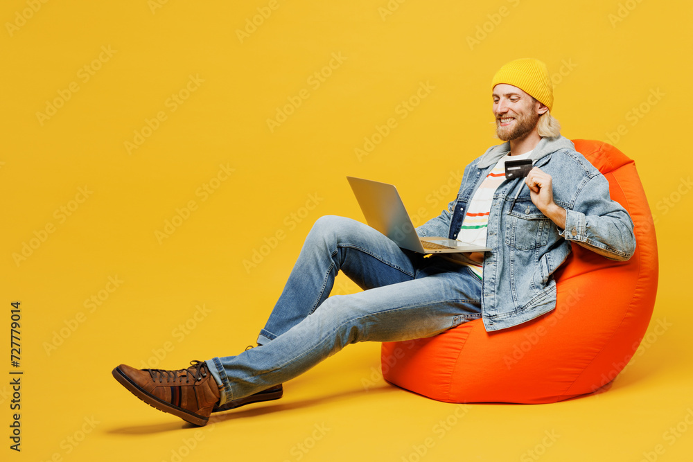 Full body young IT man he wears denim shirt hoody beanie hat casual clothes sit in bag chair hold credit bank card shopping online use work on laptop pc computer isolated on plain yellow background.