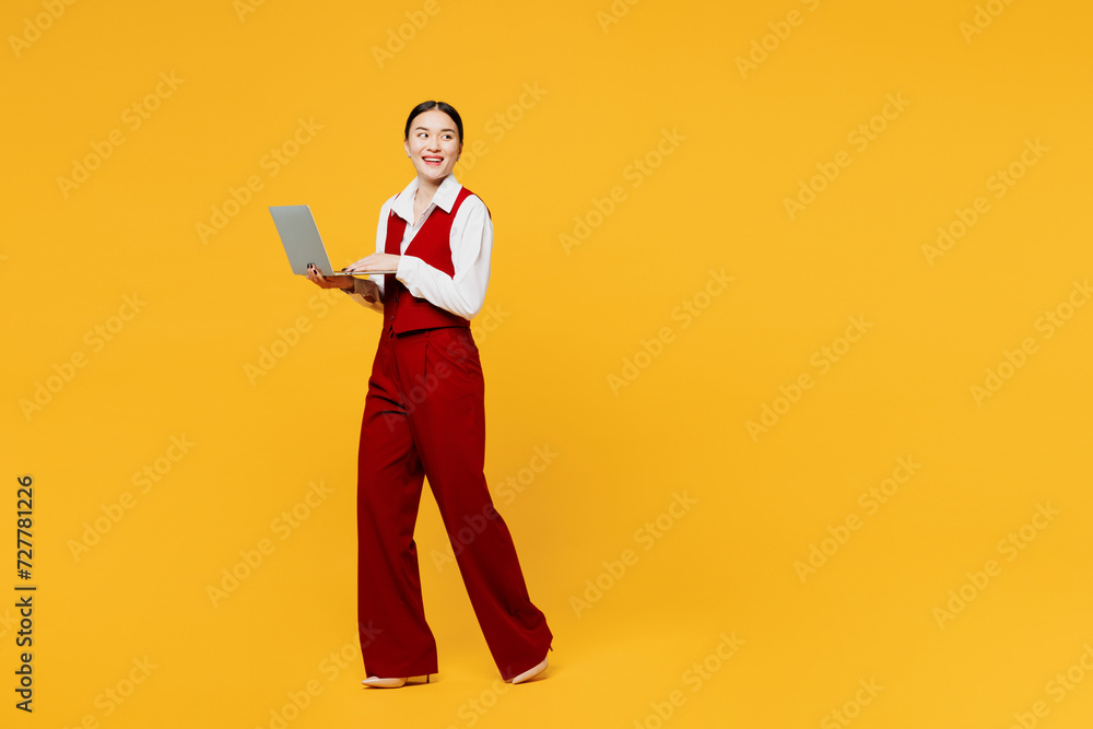 Full body side view young lawyer employee IT business woman of Asian ethnicity wear red vest shirt work at office walk go use laptop pc computer look aside isolated on plain yellow background studio. Full body side view young lawyer employee IT business woman of Asian ethnicity wear red vest shirt work at office walk go use laptop pc computer look aside isolated on plain yellow background studio.