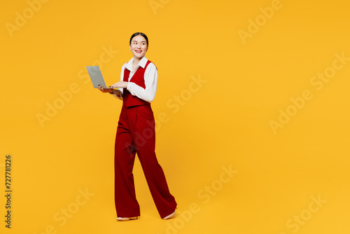 Full body side view young lawyer employee IT business woman of Asian ethnicity wear red vest shirt work at office walk go use laptop pc computer look aside isolated on plain yellow background studio.