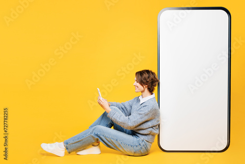 Full body young woman wears grey knitted sweater shirt casual clothes sit near big huge blank screen mobile cell phone with area use smartphone isolated on plain yellow background. Lifestyle concept.
