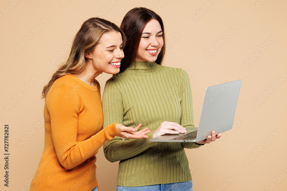 Side view young friends two IT women they wear orange green shirt casual clothes together hold use work on laptop pc computer isolated on plain pastel light beige background studio. Lifestyle concept. Side view young friends two IT women they wear orange green shirt casual clothes together hold use work on laptop pc computer isolated on plain pastel light beige background studio. Lifestyle concept.