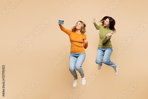 Full body young friends two women they wear orange green shirt casual clothes jump high do selfie shot on mobile cell phone post photo on social network show v-sign isolated on plain beige background.