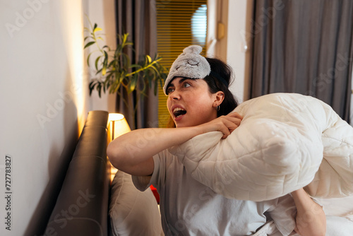Quadro em tela Portrait of annoyance angry Caucasian woman wearing sleep mask knocking with pillow to wall sitting in bed