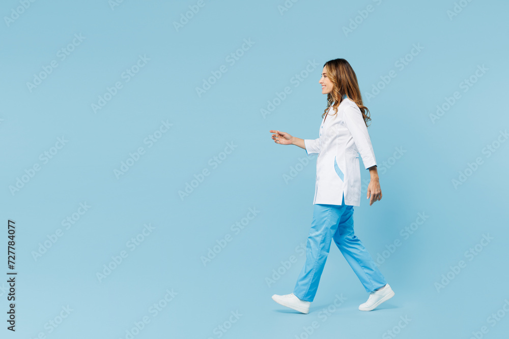 Full body side profile view smiling female doctor woman she wears white medical gown suit work in hospital clinic office walk go isolated on plain blue background studio. Health care medicine concept. Full body side profile view smiling female doctor woman she wears white medical gown suit work in hospital clinic office walk go isolated on plain blue background studio. Health care medicine concept.