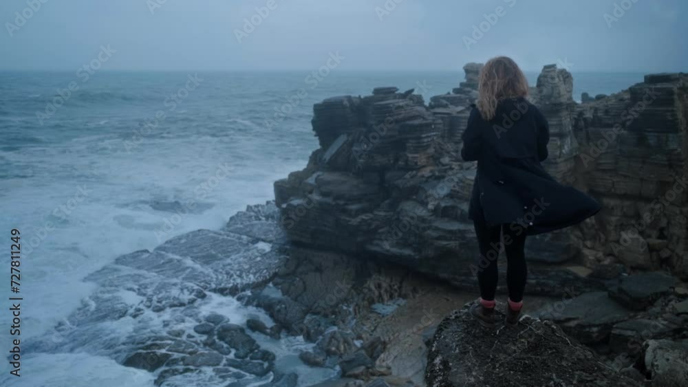 Breathtaking view of young woman stands on the edge of a cliff in black waving coat. Girl alone watching ocean waves crashing on the shore and turn into white foam. Moody atmosphere. Sadness concept Breathtaking view of young woman stands on the edge of a cliff in black waving coat. Girl alone watching ocean waves crashing on the shore and turn into white foam. Moody atmosphere. Sadness concept