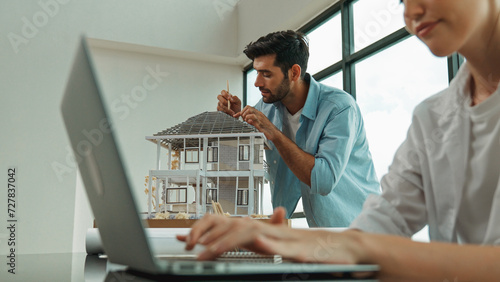 Engineer measure house model and tell cooperative coworker about house structure while coworker using laptop analysis data. Smart interior team working together at office with skyscraper. Tracery