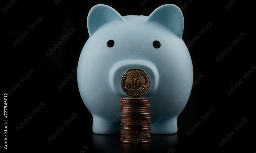 Coin in denomination of 10 Ukrainian hryvnias and a blue piggy bank against a black background - obrazy, fototapety, plakaty 