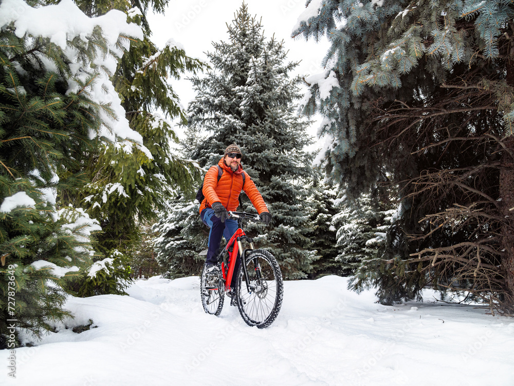 A guy rides a bicycle in a winter park among snow-covered fir trees. A cyclist in an orange jacket joyfully rides along a snowy path. Active lifestyle in winter A guy rides a bicycle in a winter park among snow-covered fir trees. A cyclist in an orange jacket joyfully rides along a snowy path. Active lifestyle in winter