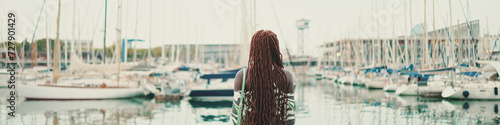 Woman with African braids wearing top looks at the yachts and ships standing on the pier in the port, Panorama, Back view.