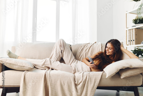 Happy woman holding mobile phone and smiling while using it on the cozy sofa in her living room